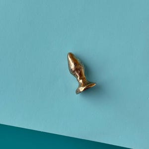 Op de afbeelding: Een kleine, goudkleurige anale plug met een afgeronde bovenkant en een uitlopende basis. Het object bevindt zich op een lichtblauwe ondergrond, met een donkerdere blauwgroene ondergrond in de linker benedenhoek.
