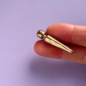 May include: A gold-colored, tapered earring with a rounded top. The earring is held against a light purple background. The earring has a smooth, polished surface and a pointed end.
