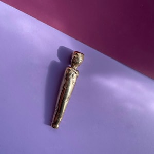 May include: A gold-colored metal figurine of a person standing upright with a rounded head and a smooth, slightly textured surface.