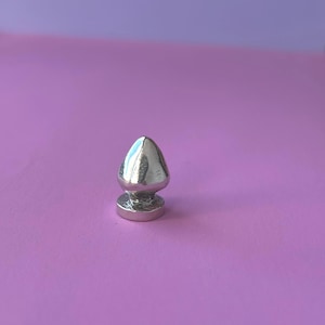 May include: A silver-coloured, metallic butt plug with a rounded base and a conical top. The product is set against a solid, light purple background. The plug has a smooth, reflective surface.