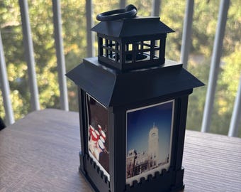 May include: A black square lantern with a tiered roof and a top hanging ring. The four sides feature inset rectangular photo panels, including one showing a tall clock tower building and another displaying a group of red and white circular objects.