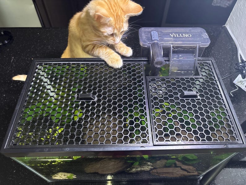 Top-down view of a fish tank with a black 3D-printed honeycomb mesh aquarium lid cover installed, with an orange cat curiously interacting with the tank. The mesh lid provides airflow and light penetration while preventing fish from jumping and pets from accessing the tank. Features two built-in feeding ports and fits around a hang-on-back filter. A stylish and protective solution for aquarium owners with cats or other curious pets.