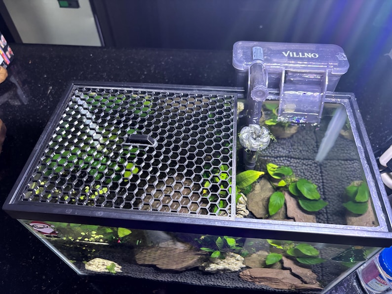 May include: A rectangular aquarium with a black frame, featuring a honeycomb-patterned lid and a filter labeled "VILLNO." The tank is filled with water, aquatic plants, rocks, and a dark substrate. The filter is positioned on the right side of the tank.