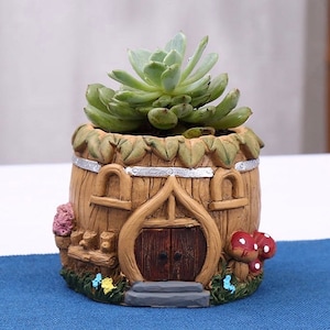 May include: A small, brown, wooden-looking planter shaped like a house with a door and windows. The planter has a green succulent plant growing inside. The planter has a silver trim around the top and a gray base.