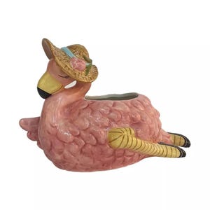 May include: Pink ceramic flamingo planter wearing a straw hat with a blue ribbon and pink flower. The flamingo has yellow legs and a yellow beak with a black tip. The planter is designed to hold small plants or flowers.