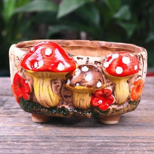 May include: A ceramic planter with a whimsical mushroom design. The planter features red and brown mushrooms with white spots, surrounded by red and orange flowers. The planter is beige with a textured surface and sits on three small feet.