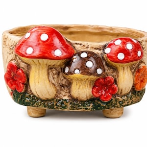 May include: A beige ceramic planter with a whimsical design featuring three mushrooms with red caps and white spots, and a brown mushroom. Red and orange flowers and green grass add color to the planter.