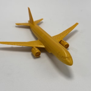 May include: A small, mustard-yellow 3D-printed model airplane featuring a fuselage, two wings, a vertical stabilizer, and two engine nacelles mounted under the wings. The surface shows fine horizontal layer lines from the printing process.