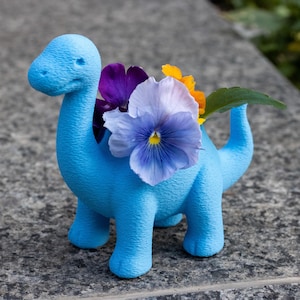 Puede incluir: Una maceta con forma de dinosaurio azul claro llena de flores coloridas. La maceta tiene una superficie texturizada y está sobre una superficie de piedra gris. Las flores incluyen pensamientos morados, blancos y amarillos.