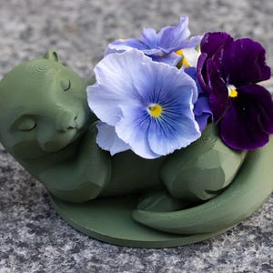 May include: A green otter-shaped planter holding a cluster of colorful pansies. The flowers include shades of blue, purple, and yellow. The otter is lying down, with its eyes closed, and is resting on a circular base.