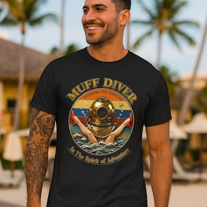 May include: Black t-shirt with a vintage-style graphic featuring a diving helmet, red high heels, and the text "Muff Diver" and "In The Spirit of Adventure". The design is set against a retro sunset background.