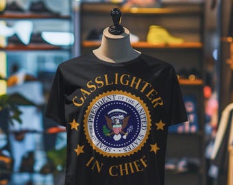 May include: Black t-shirt with the words "Gasslighter in Chief" in gold lettering. The shirt features a circular design with the presidential seal in the center, surrounded by stars. The shirt is displayed on a mannequin.