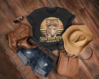 May include: Black t-shirt with a graphic of a cat wearing sunglasses in front of a sunset, with the text "BOW DOWN TO MY PEASANTS". The shirt is paired with denim shorts, a brown belt, cowboy boots, a straw hat, sunglasses, and a brown purse.
