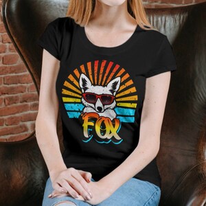 Peut inclure: Un t-shirt noir avec un graphique r&eacute;tro d'un renard portant des lunettes de soleil. Le graphique pr&eacute;sente un coucher de soleil avec des rayures orange, jaunes, bleues et blanches. Le mot "FOX" est &eacute;crit dans une police stylis&eacute;e sous le renard.