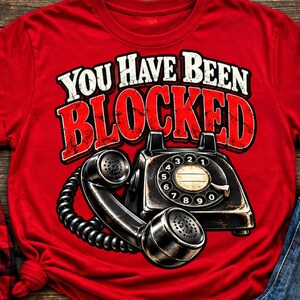 May include: A bright red t-shirt featuring a graphic of a black vintage rotary telephone with a coiled cord. Above the telephone, the text reads "YOU HAVE BEEN BLOCKED" in bold white and red distressed lettering with a black outline.