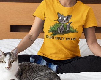 May include: Mustard yellow t-shirt featuring a cartoon raccoon sitting in grass. The raccoon is gray and white with a striped tail. The text "DON'T SNACK ON ME" is below the raccoon. A cat is in the foreground.