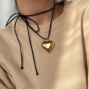 Op de afbeelding: Een gouden hartvormige hangerketting met een zwarte choker-stijl ketting. De ketting wordt gedragen door een persoon met een lichtbruin shirt.