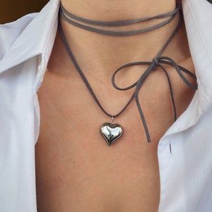 Può includere: Una collana con un ciondolo a forma di cuore argentato e un choker in pelle scamosciata grigia. Il choker è legato con un fiocco sul retro del collo.