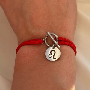Op de afbeelding: Rode koordarmband met een zilverkleurige sluiting en een ronde bedel. De bedel toont het sterrenbeeld Leeuw. De armband wordt om de pols gedragen.