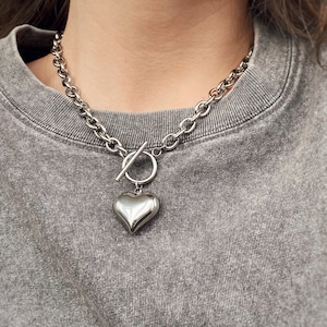 May include: A silver chain necklace with a large, heart-shaped pendant. The chain is thick and chunky, and the pendant is smooth and shiny.