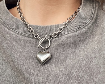 May include: A silver chain necklace with a large, heart-shaped pendant. The chain is thick and chunky, and the pendant is smooth and shiny.