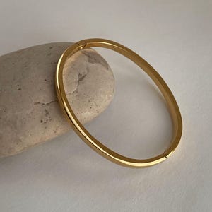 Peut inclure: Un bracelet jonc dor&eacute; &agrave; l'aspect simple et arrondi. Le bracelet est en m&eacute;tal et a une finition lisse.