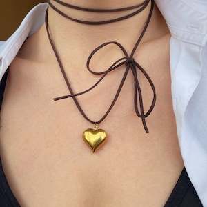 May include: A gold heart pendant necklace on a brown leather cord. The cord is tied in a bow at the back of the neck.