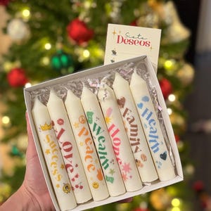 May include: A box of seven white candles with colorful text and illustrations. Each candle has a different word: Success, Love, Peace, Wealth, Hope, Family, and Health. A small card with the text "Siete Deseos" is also in the box.