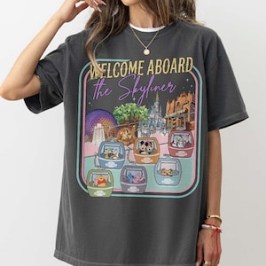 May include: Dark gray t-shirt with a colorful graphic featuring the text "WELCOME ABOARD the Skyliner" and images of Disney characters in gondolas. The graphic includes a park scene with a purple sphere and castle.