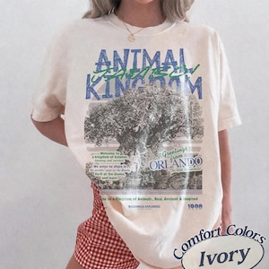 May include: An ivory short-sleeve t-shirt featuring a large graphic print with the text ANIMAL KINGDOM, Greetings from ORLANDO, and Wilderness Explorers. The design includes a detailed illustration of a large tree and various text elements in blue and green ink, printed on a light-colored cotton fabric.