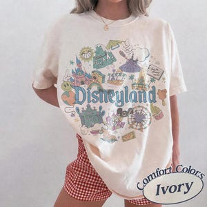 May include: Ivory-colored t-shirt with a Disneyland graphic. The graphic features various park icons, including a castle, a Ferris wheel, and the Disneyland logo in blue. The shirt is labeled "Comfort Colors" and "Ivory".