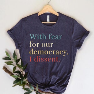 Può includere: T-shirt blu navy con la scritta "With fear for our democracy, I dissent." in turchese, giallo e rosso. La maglietta è appesa a una gruccia di legno, con un rametto di verde nell'angolo.