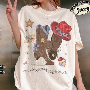 Pu&ograve; includere: Una t-shirt color avorio con una stampa grafica di stivali da cowboy marroni, un cappello da cowboy rosso, una stella da sceriffo, palloncini e il testo TAY STORY. Il design include piccoli fiori, stelle e una corda di perline colorata.