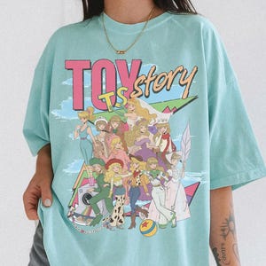 Peut inclure: Un t-shirt bleu clair avec une grande impression graphique portant le texte "TOY story" en rose et jaune. Le motif pr&eacute;sente un groupe de personnages f&eacute;minins illustr&eacute;s dans diverses tenues, incluant une balle jaune et des formes g&eacute;om&eacute;triques.
