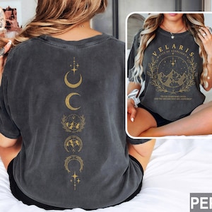 May include: Dark gray t-shirt with a gold design featuring phases of the moon on the back and the text "VELARIS" with a mountain and star design on the front. The shirt is a relaxed fit.