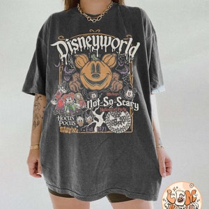 Op de afbeelding: Een zwart T-shirt met een vintage-stijl grafische afbeelding van Mickey Mouse en zijn vrienden die Halloween vieren in Disney World. De afbeelding bevat een pompoen-gekopte Mickey Mouse, een griezelige boom en de tekst "Mickey's Not-So-Scary Halloween Party" en "Est. 1971".