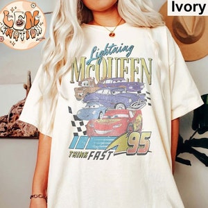 May include: A white t-shirt with a vintage-style graphic of Lightning McQueen from the Cars movie. The graphic features the text "Lightning McQueen" and "Think Fast" along with a checkered flag and the number 95. The shirt is a light ivory color.