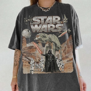 Può includere: T-shirt grafica di Star Wars grigio scuro con Darth Vader, camminatori AT-AT e caccia X-wing. Il logo di Star Wars è stampato in alto. La maglietta ha un aspetto vintage e usurato.