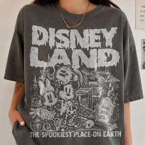 May include: Charcoal grey t-shirt with a distressed "DISNEY LAND" graphic. The graphic features Mickey and Minnie Mouse skeletons in front of a spooky scene, with the text "THE SPOOKIEST PLACE ON EARTH".