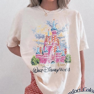 May include: Ivory-colored t-shirt featuring a colorful graphic of a pink castle with blue turrets, green bushes, and the text "This is Now! Walt Disney World 1997." The shirt is from Comfort Colors.