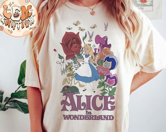 May include: A white t-shirt with a graphic print of Alice in Wonderland. The graphic features Alice standing in a garden with a large red rose, flowers, and other characters from the story. The text "Alice in Wonderland" is printed below the graphic.