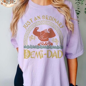 以下が含まれることがあります： 「Just an Ordinary Demi-Dad」というテキストと、漫画のキャラクターのグラフィックが描かれた紫色のTシャツです。