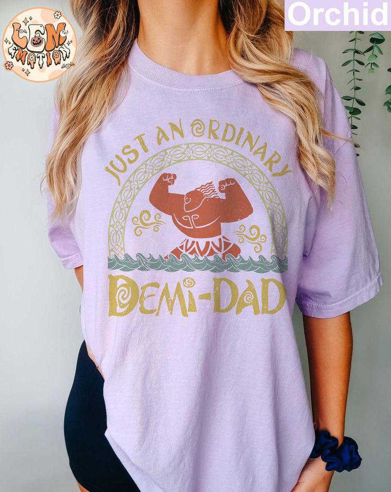 以下が含まれることがあります： 「Just an Ordinary Demi-Dad」というテキストと、漫画のキャラクターのグラフィックが描かれた紫色のTシャツです。