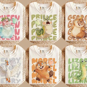 Puede incluir: Seis camisetas crema dobladas con gráficos de animales de dibujos animados y nombres. Los diseños incluyen una sirena, una oruga, un castor, un cisne y un lagarto. Cada camisa tiene un diseño y texto únicos.
