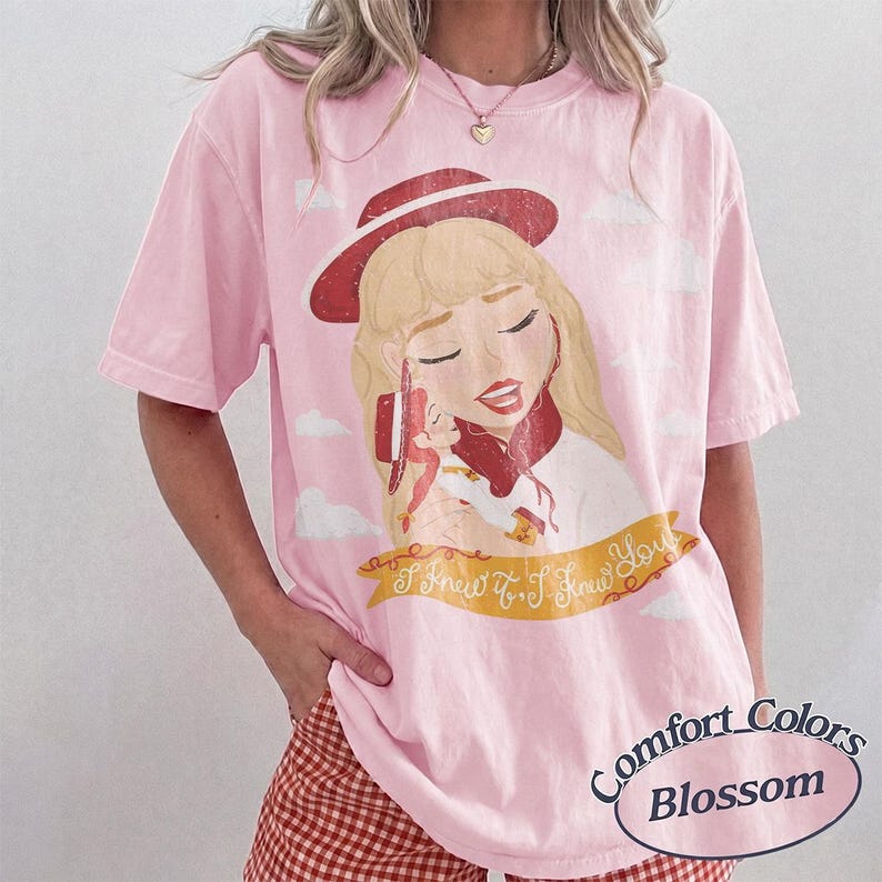 Pode incluir: Uma t-shirt de manga curta rosa claro com uma ilustra&ccedil;&atilde;o gr&aacute;fica de uma personagem loira a usar um chap&eacute;u vermelho. Abaixo da personagem encontra-se uma faixa amarela com o texto I Knew It, I Knew You. A t-shirt tem um corte descontra&iacute;do.
