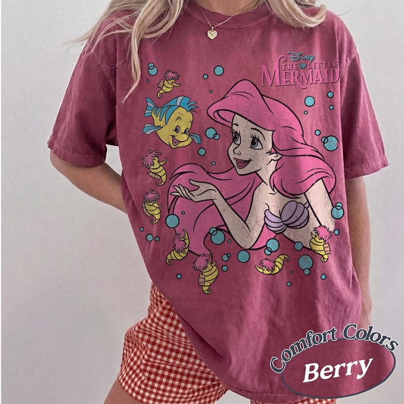 May include: A berry-colored short-sleeve t-shirt featuring a graphic of Ariel from The Little Mermaid with Flounder and several seahorses. The design includes blue bubbles and the text Disney The Little Mermaid. The shirt has a vintage-style wash.