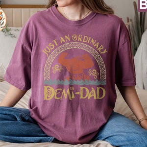 以下が含まれることがあります： ヴィンテージスタイルのフォントで「Just an Ordinary Demi-Dad」と書かれたパープルのTシャツ。テキストは、スタイリッシュな神話上の生き物のイメージが描かれた装飾的なボーダーで囲まれています。