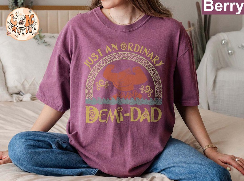以下が含まれることがあります： ヴィンテージスタイルのフォントで「Just an Ordinary Demi-Dad」と書かれたパープルのTシャツ。テキストは、スタイリッシュな神話上の生き物のイメージが描かれた装飾的なボーダーで囲まれています。