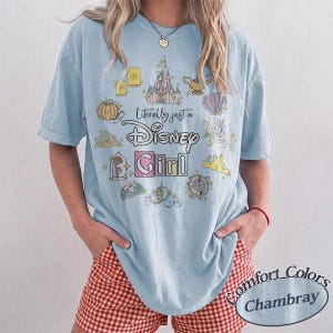 Op de afbeelding: Lichtblauw T-shirt met de tekst "Literally just a Disney Girl" en diverse Disney-thema graphics. Het shirt heeft korte mouwen en wordt gecombineerd met een rood-witte geruite short.