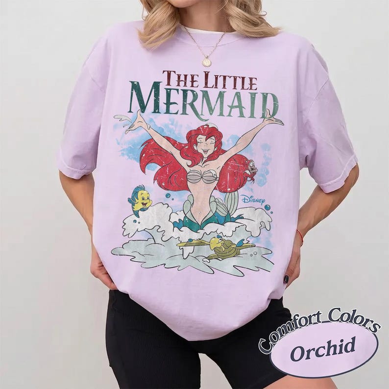 May include: A light purple short-sleeve t-shirt featuring a graphic print of Ariel from The Little Mermaid with red hair, a green tail, and the character Flounder. The text reads The Little Mermaid and Disney in a distressed, vintage-style font.